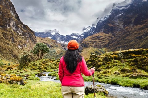 An Ecuadorian woman in a red jacket and orange hat stands by a stream, surrounded by the majestic
