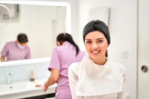 A smiling client wearing a headband and protective bib sits in a well-lit aesthetic clinic,