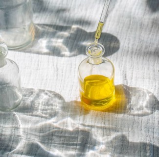A glass bottle filled with golden natural body oil sits in soft sunlight as a pipette adds more