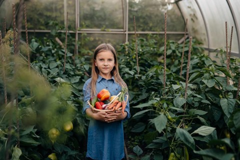 A young girl stands in a lush greenhouse filled with greenery, holding a basket of freshly picked