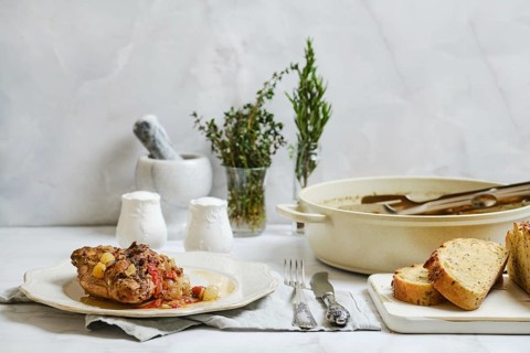 Rabbit stew on a plate next to bread slices. Small glass jars hold fresh herbs. A pot sits on the