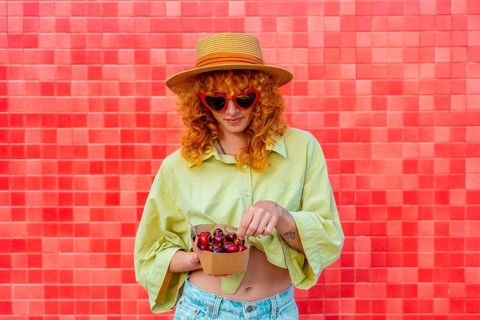 Fashionable redhead woman with heart shaped sunglasses and straw hat enjoying fresh cherries from a