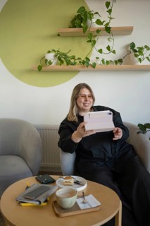 A cheerful woman relaxes in a cozy cafe lounge, interacting with a digital device. Surrounded by