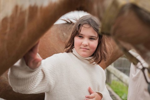 A young girl in a cozy sweater gently touches a horse on a sunny farm day. The scene captures the