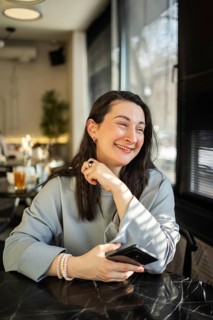 A cheerful mid-aged woman is pictured sitting comfortably at a cafe, holding a smartphone in her