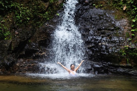 A young woman joyfully raises her arms while standing below a cascading waterfall in the lush Choco