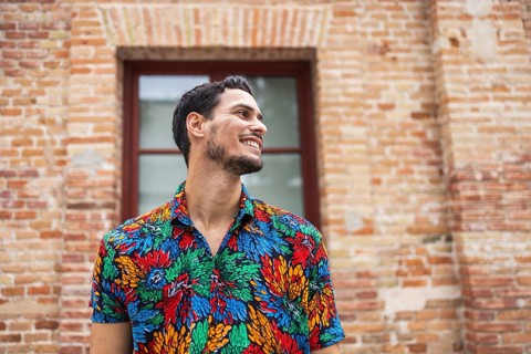 A joyful Cuban man stands against a rustic brick wall, wearing a vibrant, colorful shirt. His