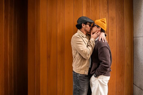 A loving Colombian gay couple shares a tender kiss while standing against a warm wooden wall. Their