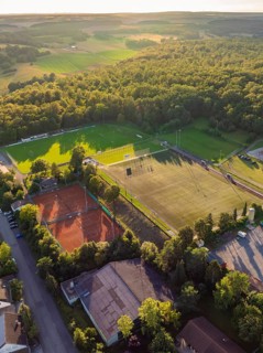 Aerial view of a sports field with football pitches and tennis courts, surrounded by woods and