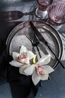 A sophisticated table setting with a textured plate, dark cutlery, and elegant white orchids. Two