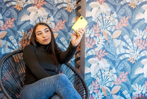 A young woman captures a selfie using her vibrant yellow mobile phone. She's seated in a stylish