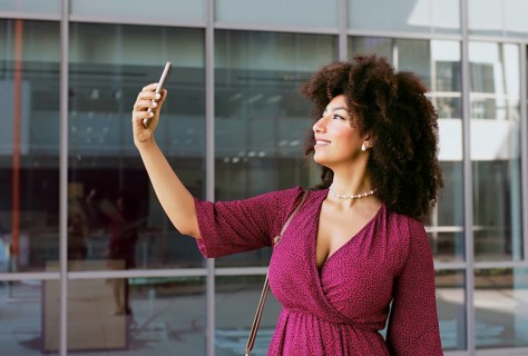 Young Arab woman with afro hair and in a stylish pink dress joyfully takes a selfie using her