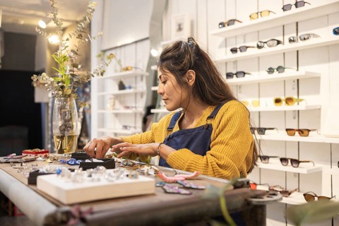 A shop assistant organizes accessories on a table in a sunglasses store. She is wearing a yellow