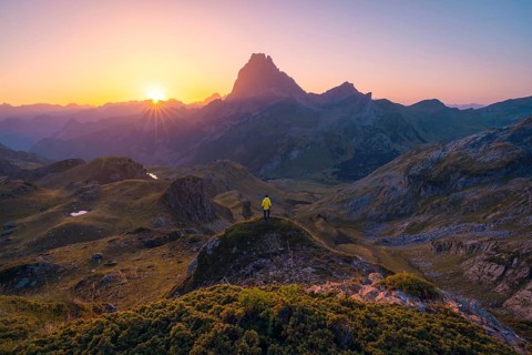 A lone hiker in a yellow jacket overlooks a mountainous landscape, bathed in the golden hues of