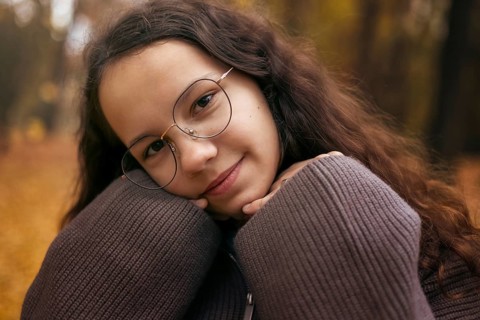 A young girl with glasses, wrapped in a cozy sweater, poses against an autumn backdrop. Her smile