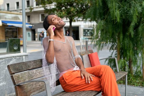 A man in vibrant fashion sits on a bench, exuding joy and pride. His mesh top and orange pants