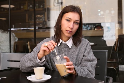 A woman in a gray sweater stirs her iced coffee while seated at an outdoor bar. She gazes