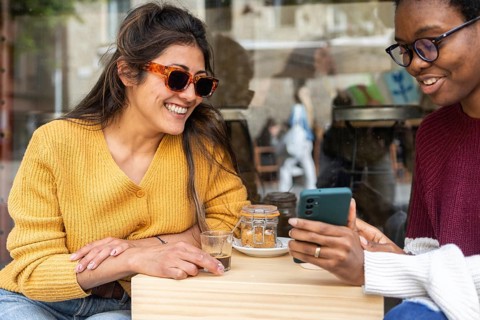 Two friends enjoy a sunny day at a cafe, sharing coffee and smiles. One wears stylish sunglasses