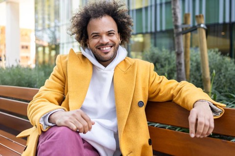 Smiling Afro man in a vibrant yellow coat enjoys a relaxed moment sitting on a bench outdoors.