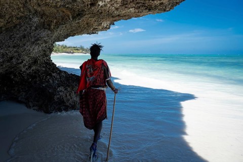 A Maasai man in traditional red clothing walks along a pristine white sand beach under a dramatic