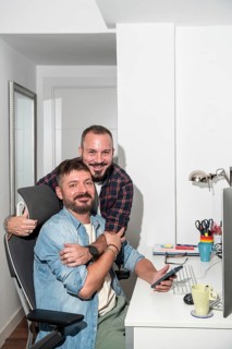 A loving gay couple is collaborating at their home office desk. One is seated, holding a
