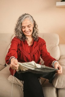 An older woman with gray hair knits contently on a beige sofa, wearing a red blouse. The cozy