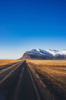 A straight dirt road stretches toward a dramatic ridge of snow-covered mountains in the Stokksnes