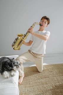 A young boy in a bright room plays a saxophone while kneeling on a carpet. His dog rests beside