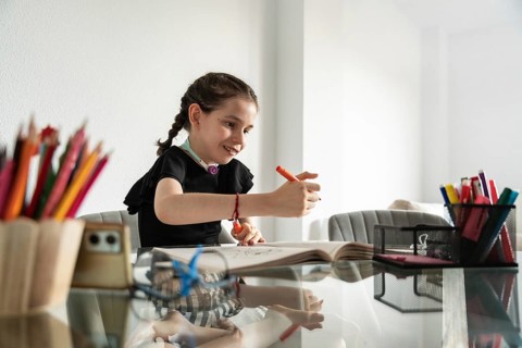 A young disabled girl with a tracheostomy happily draws at a table, surrounded by colorful pencils,