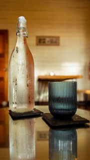 A chilled glass bottle and a textured tumbler sit on square coasters atop a reflective table. Warm,