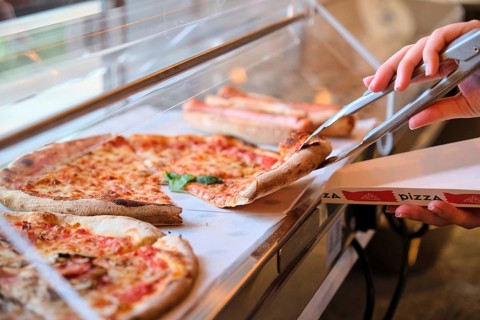 Metal tongs grabbing slice of margherita pizza from display case in pizzeria, ready to be served in