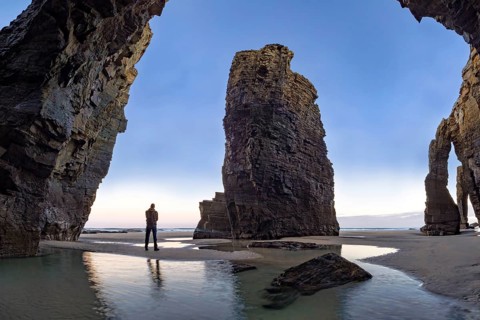 A person stands at the base of towering coastal rock formations, surrounded by reflective water