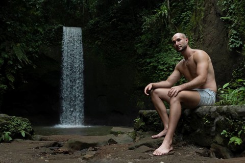 A man sits calmly by a waterfall in a lush, green forest setting in Bali. The serene scene captures