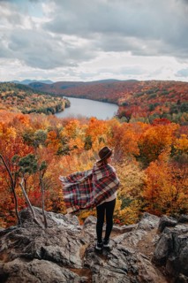 A person stands on a rocky outcrop overlooking a serene lake surrounded by vibrant autumn foliage