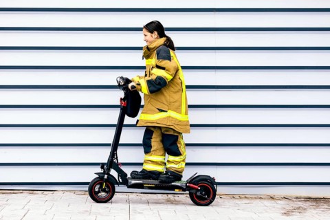 A young girl dressed in a firefighter suit rides an electric scooter in front of a striped wall,