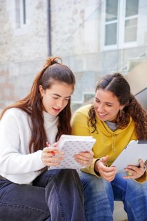 Two students sit on stairs, engaged in study and discussion One takes notes in a notebook, while