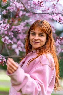 Young woman with long red hair wearing pink hoodie gently touching cherry blossoms on a spring day