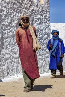 Two men in traditional Tuareg dress walk in front of a white plastered wall, local people in the