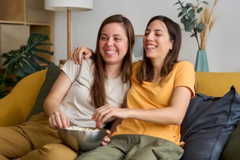 A happy lesbian couple sits comfortably on the couch, sharing a bowl of popcorn and laughing