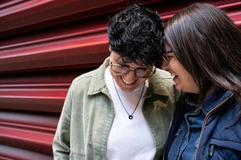 A joyful lesbian couple shares a laugh against a bright red backdrop. Their affectionate embrace