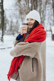 A cheerful young woman wrapped in a cozy coat and a red scarf, holding a coffee cup in a snowy park