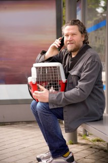 A man sits at a bus stop holding a cat carrier, engaged in conversation on his phone. The scene