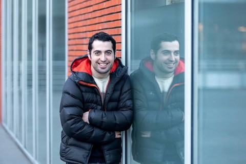 A man in a warm jacket leans against a glass wall, his reflection visible beside him. The setting