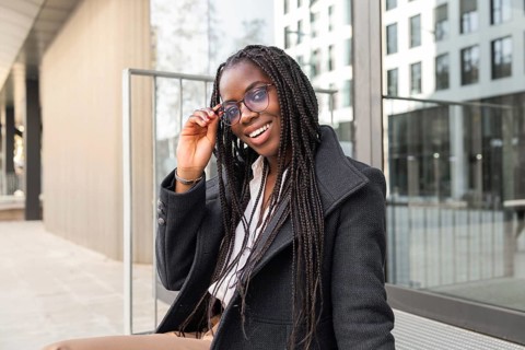 A young African American businesswoman with braided hair enjoys a break outdoors, smiling while