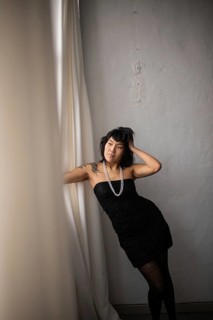 A graceful Asian woman poses by a textured wall, wearing a strapless black dress and pearl necklace