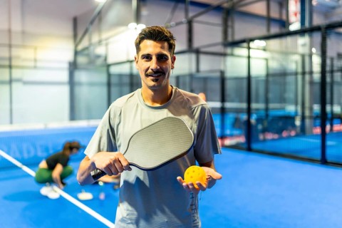 Young male athlete standing on a bright blue indoor pickleball court, holding a paddle and a yellow