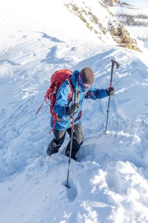 A skier in winter gear climbs a snow-covered mountain with poles and a backpack. The bright winter