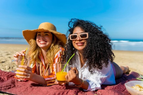 Two smiling young women lying on a blanket at the beach, enjoying refreshing orange drinks with