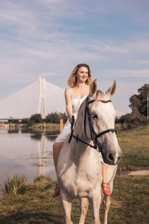 A young woman in a white dress rides a white horse beside a shimmering river. A striking cable