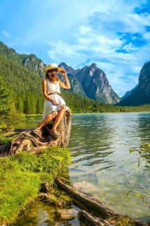 Young woman with straw hat sitting on a tree trunk by the lake admiring the mountain landscape in